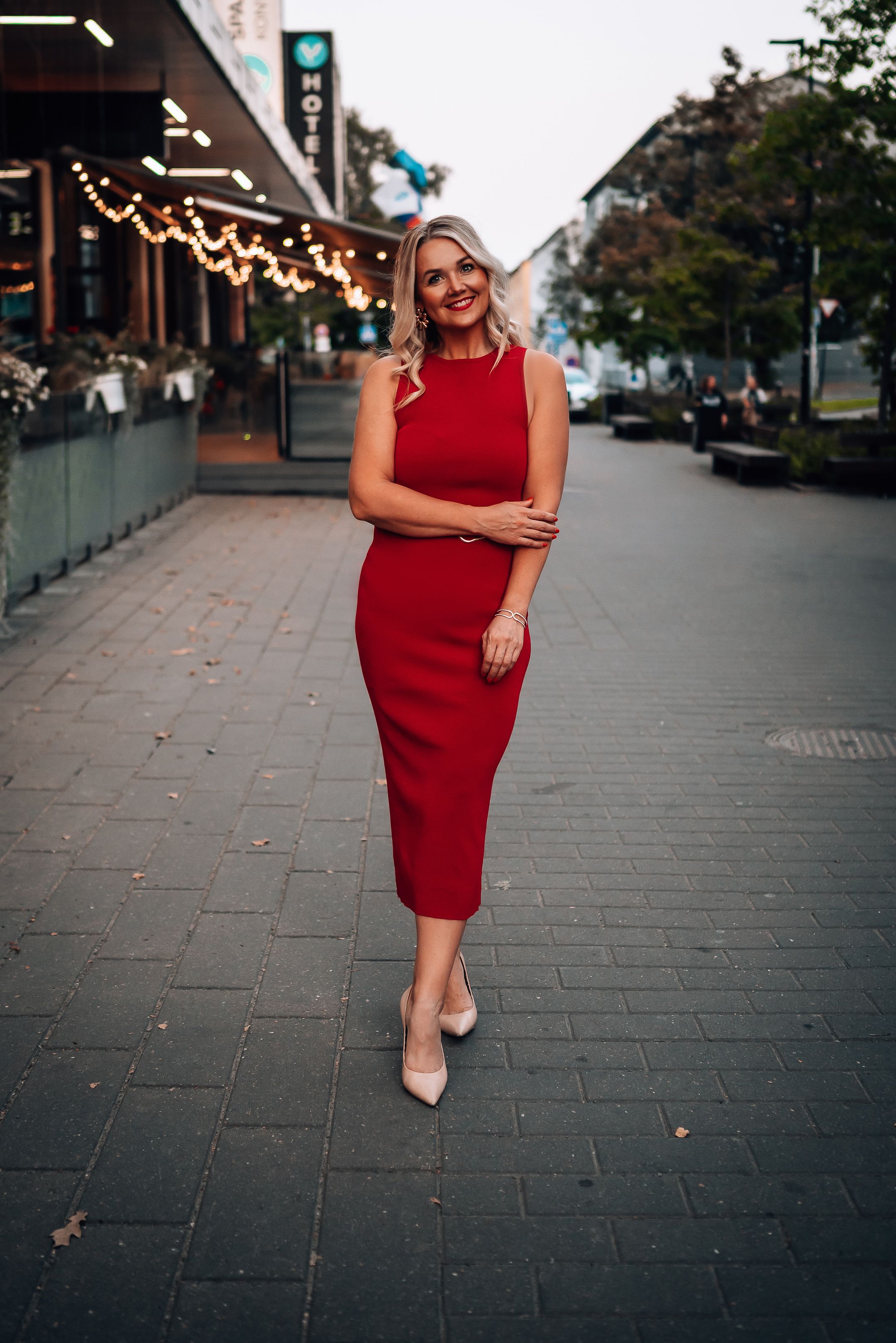Woman in red dress poses confidently on urban sidewalk with string lights and buildings in background at dusk.