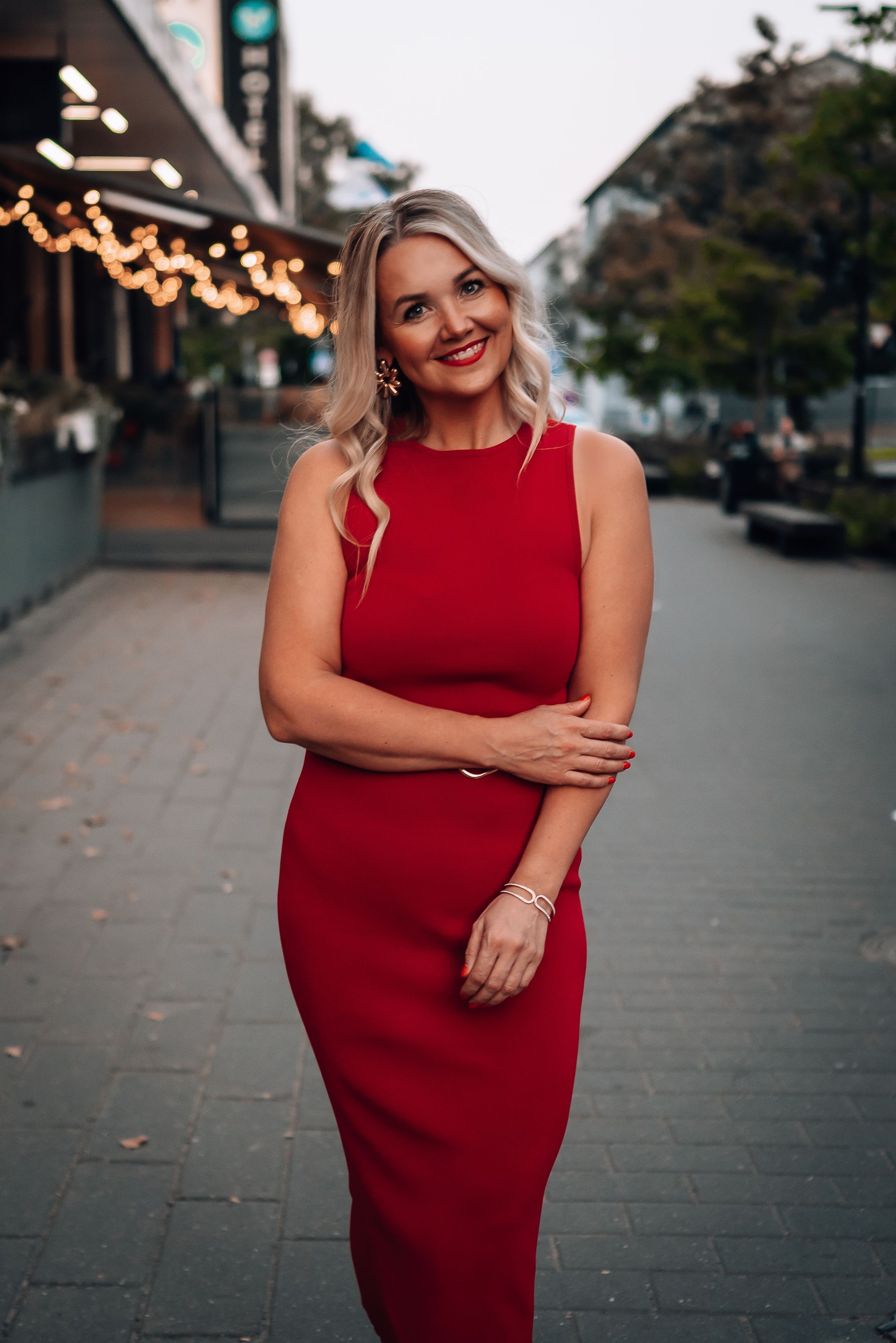 A woman in a sleeveless red dress poses confidently on a city street with string lights in the background.