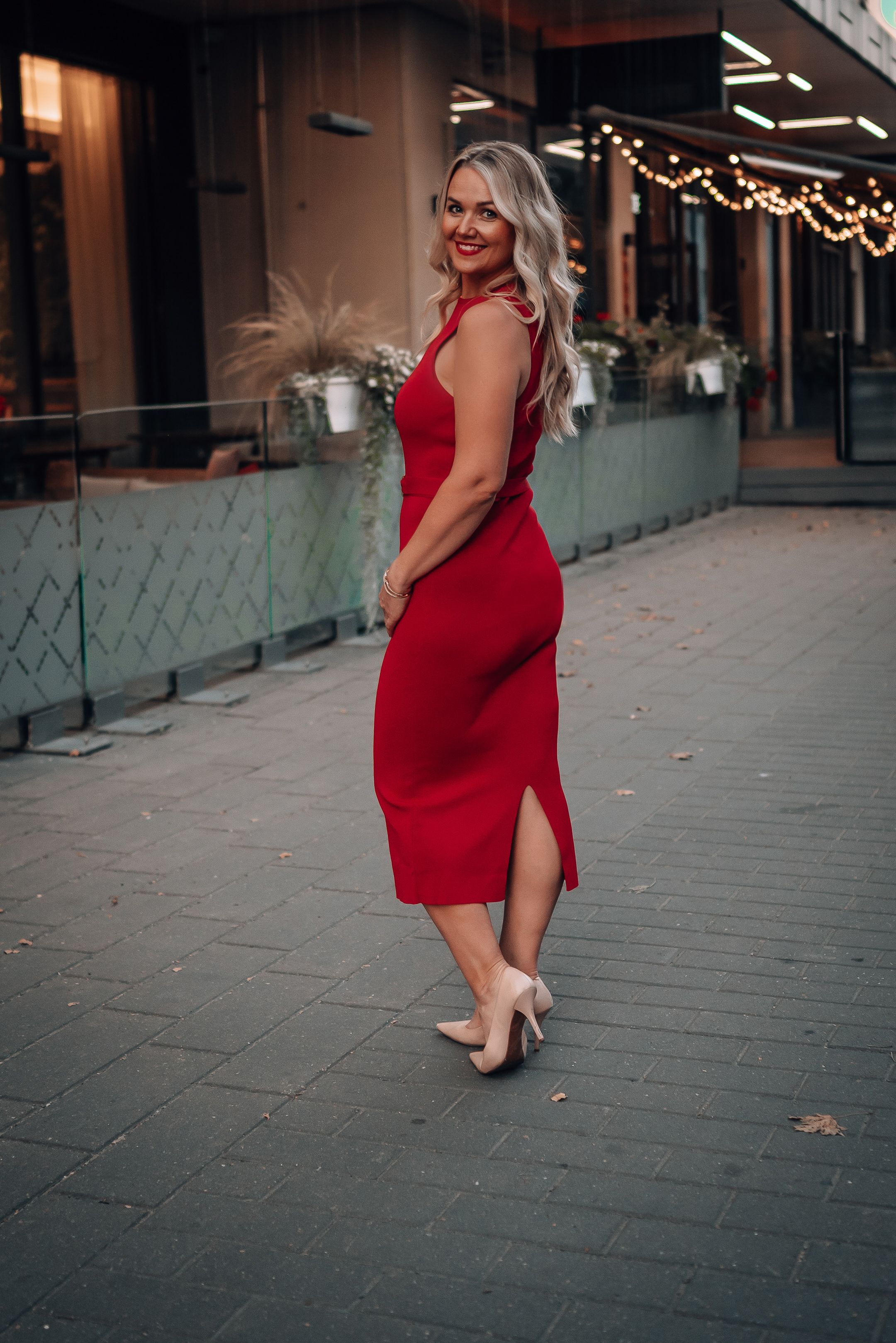 A woman in a fitted red dress stands elegantly on a brick sidewalk near illuminated storefronts.