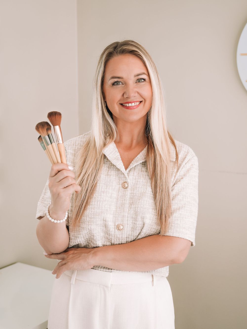 Smiling blonde makeup artist holding professional brushes in a bright studio wearing a white textured blouse.