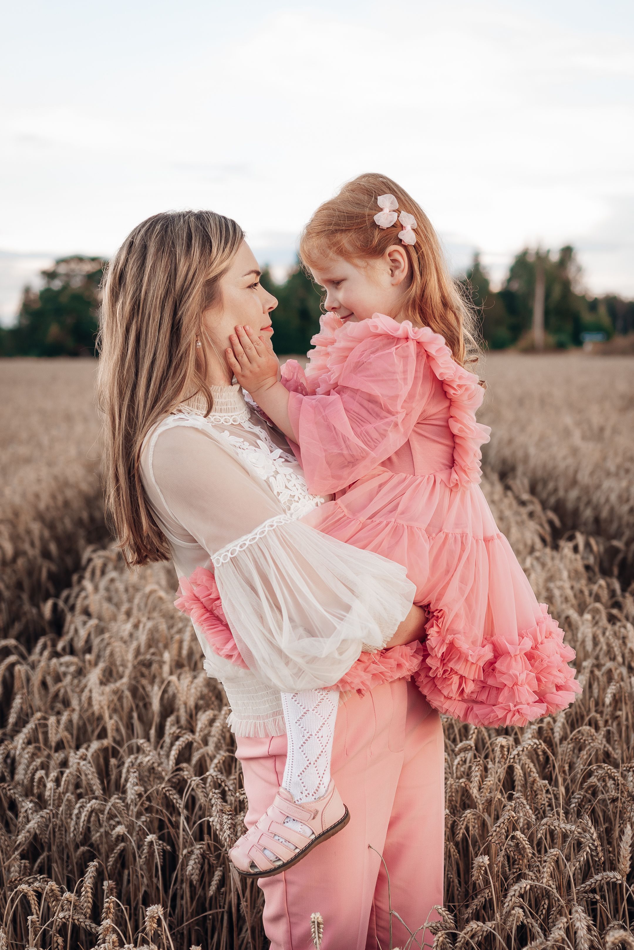A tender moment between parent and child wearing pink and white dresses while standing in a wheat field at sunset.