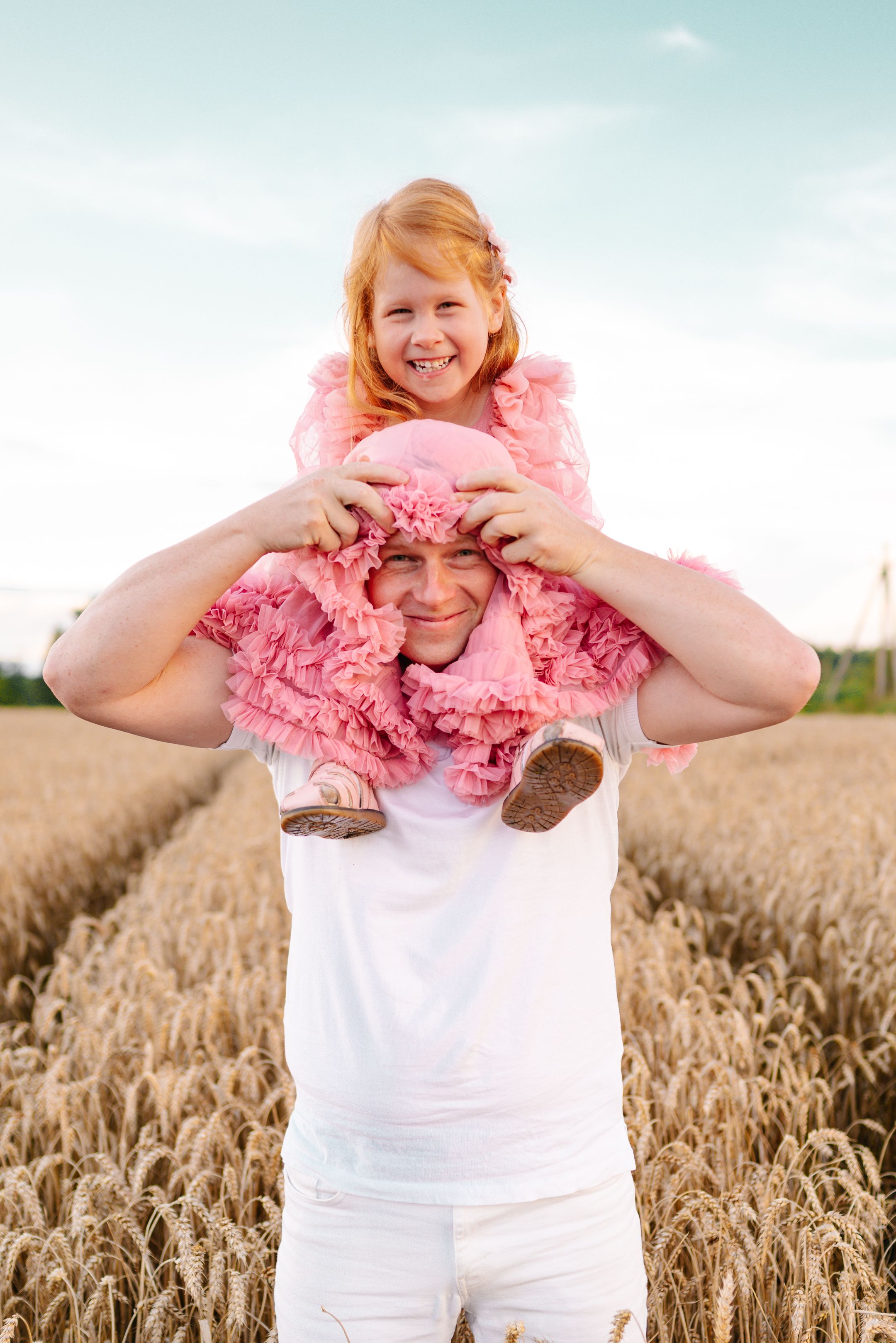 Playful scene in a golden wheat field with someone wearing pink ruffled clothing sitting on shoulders at sunset.