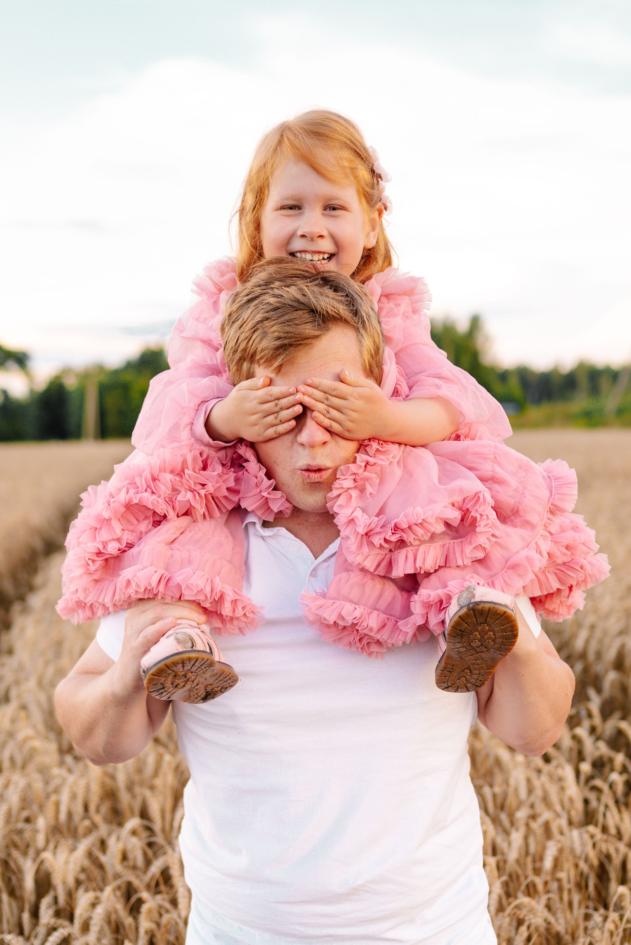 Child wearing white shirt and pink tulle plays peek-a-boo in golden wheat field during sunset.