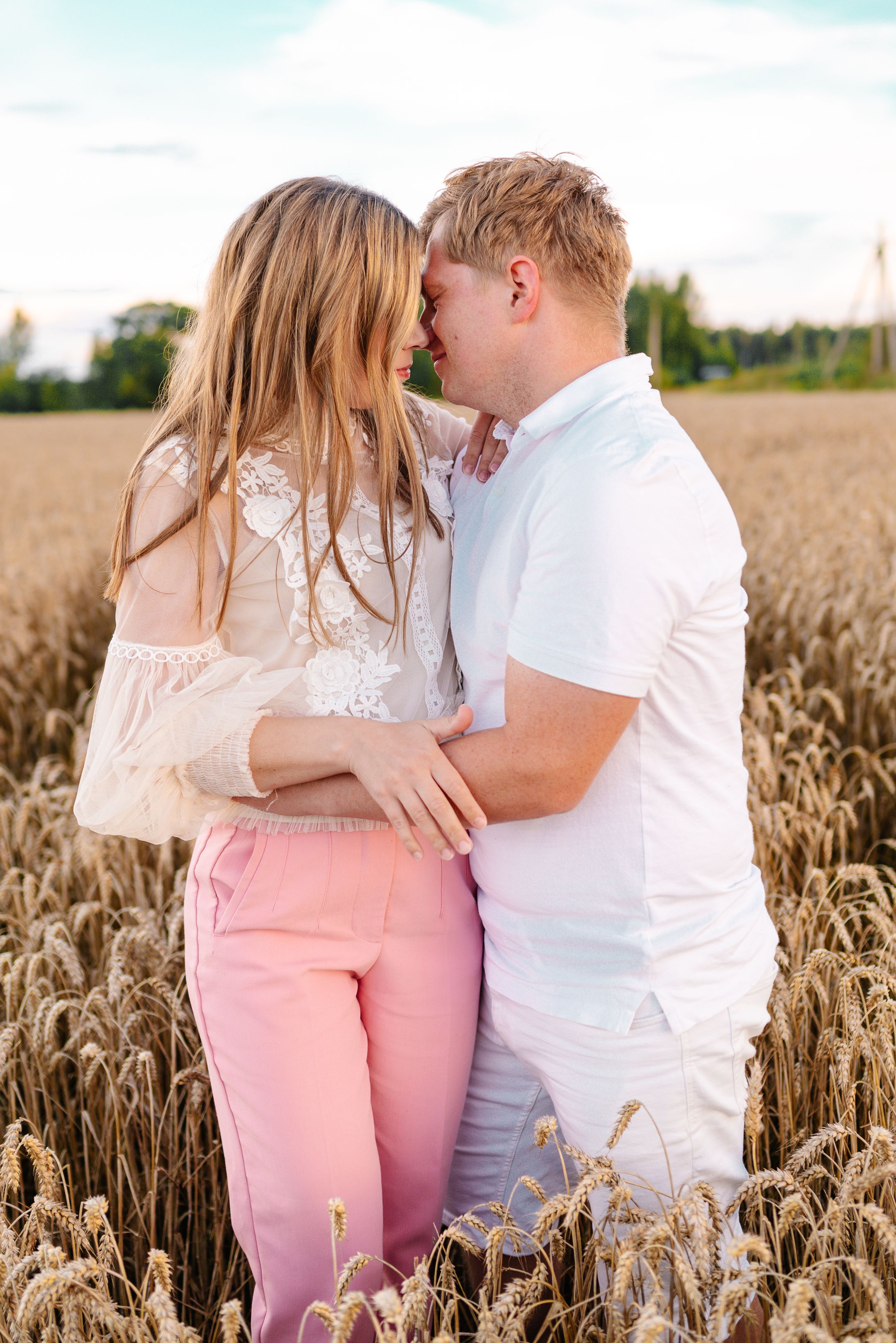 A couple embraces intimately in a golden wheat field during sunset, wearing white and pink casual clothing.