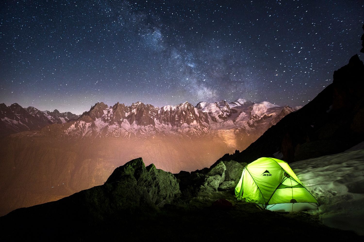 Glowing green tent under a stunning Milky Way sky above snow-capped mountain peaks.