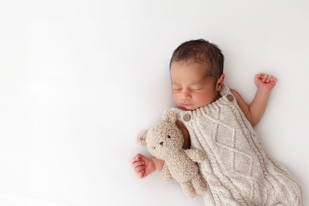 Little newborn boy sleeps on his back with a teddy bear that matches his knitted romper
