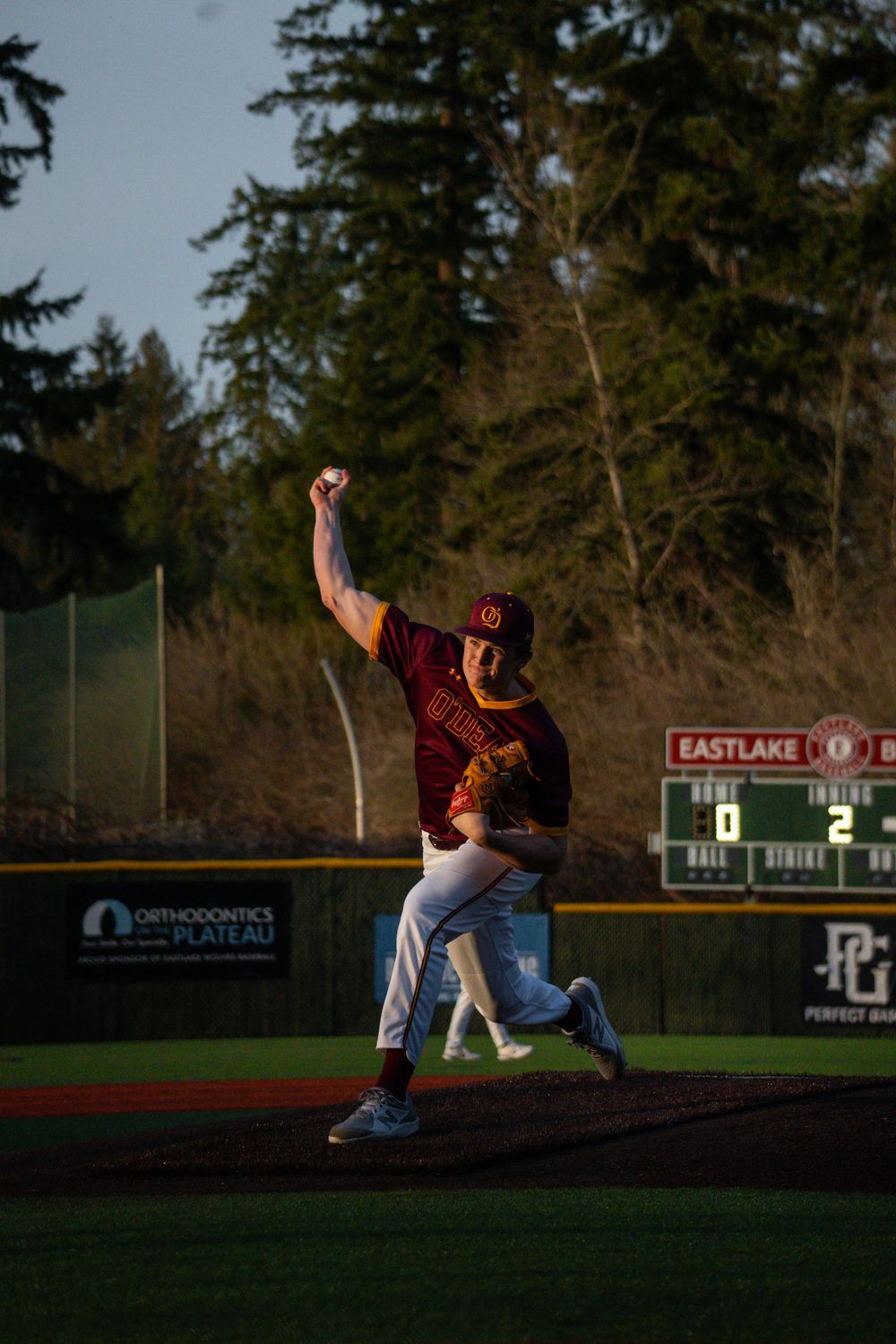 Baseball pitcher in maroon uniform throws from the mound during golden hour at a field with evergreen trees in background.
