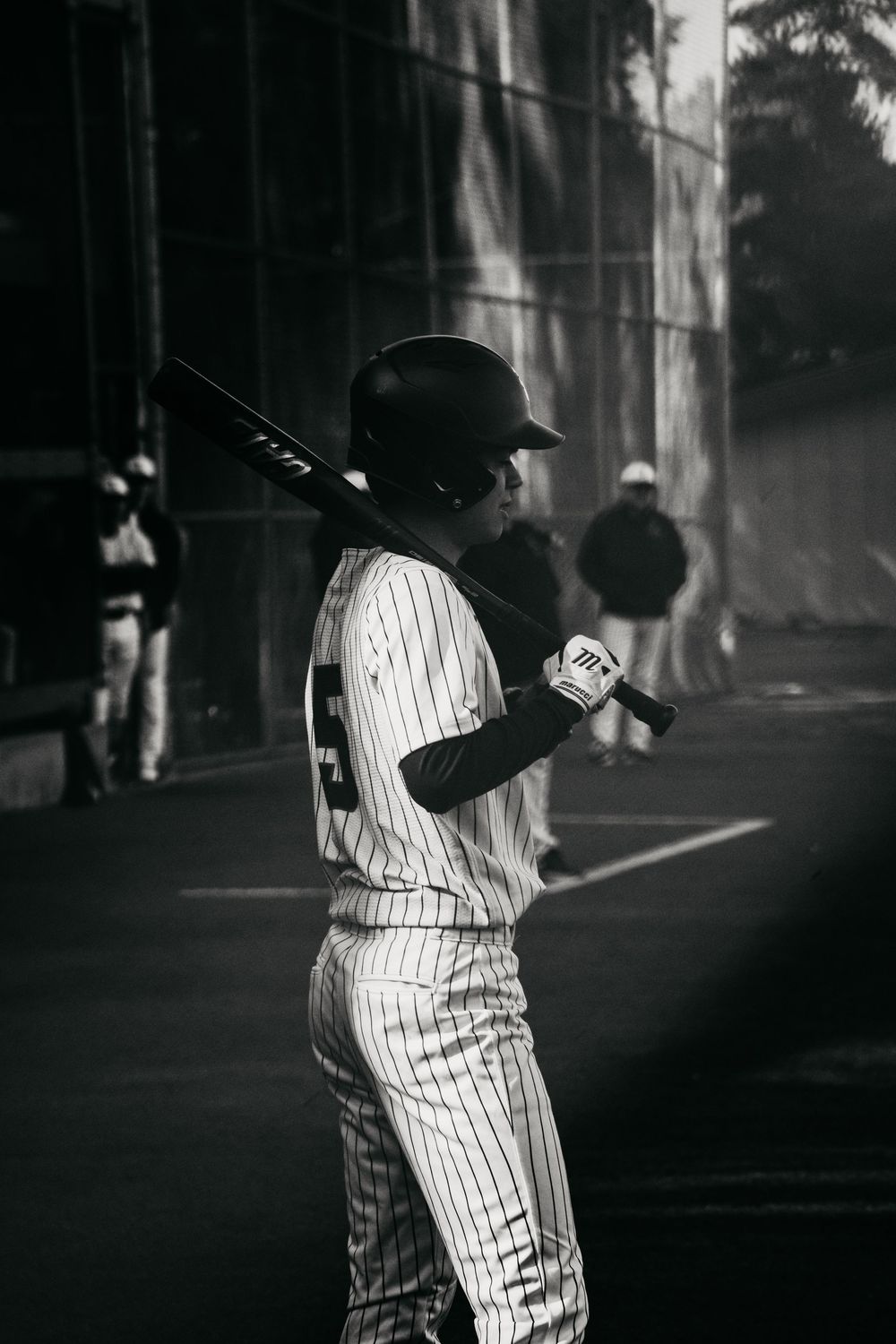 A baseball player in pinstripe uniform stands in dramatic black and white lighting at a baseball field.