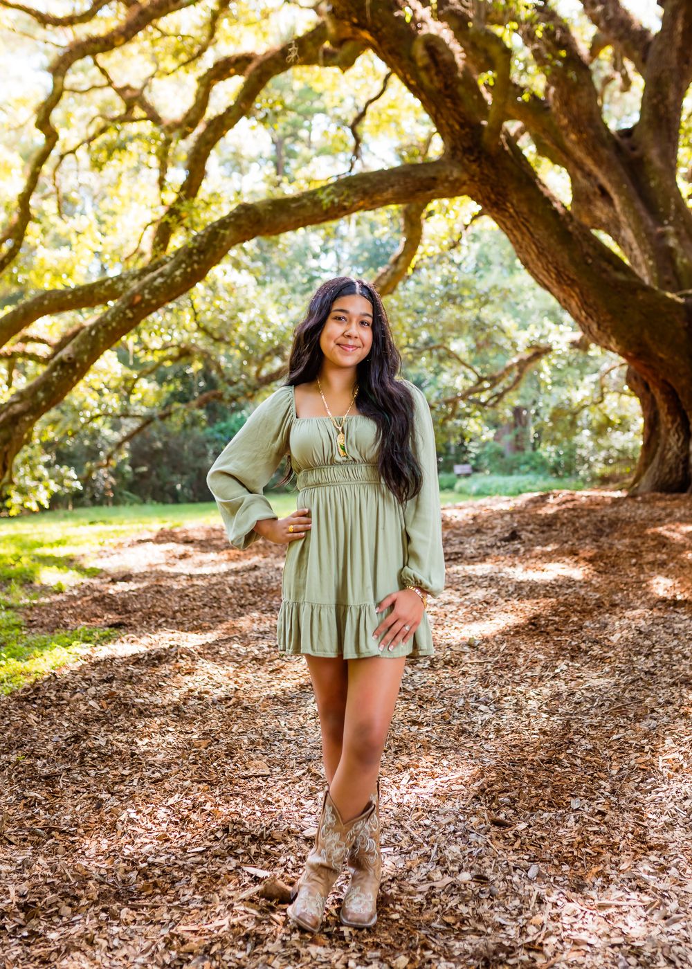 Woman in sage green dress and brown boots poses under majestic oak trees in a natural outdoor setting.