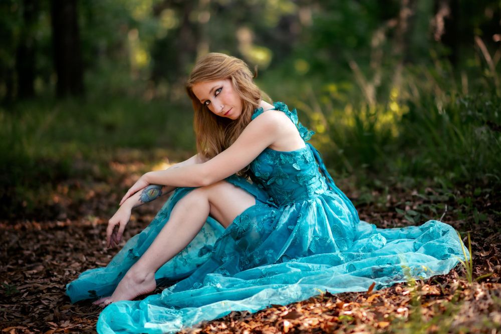 Model in turquoise gown sits gracefully on forest floor surrounded by greenery.