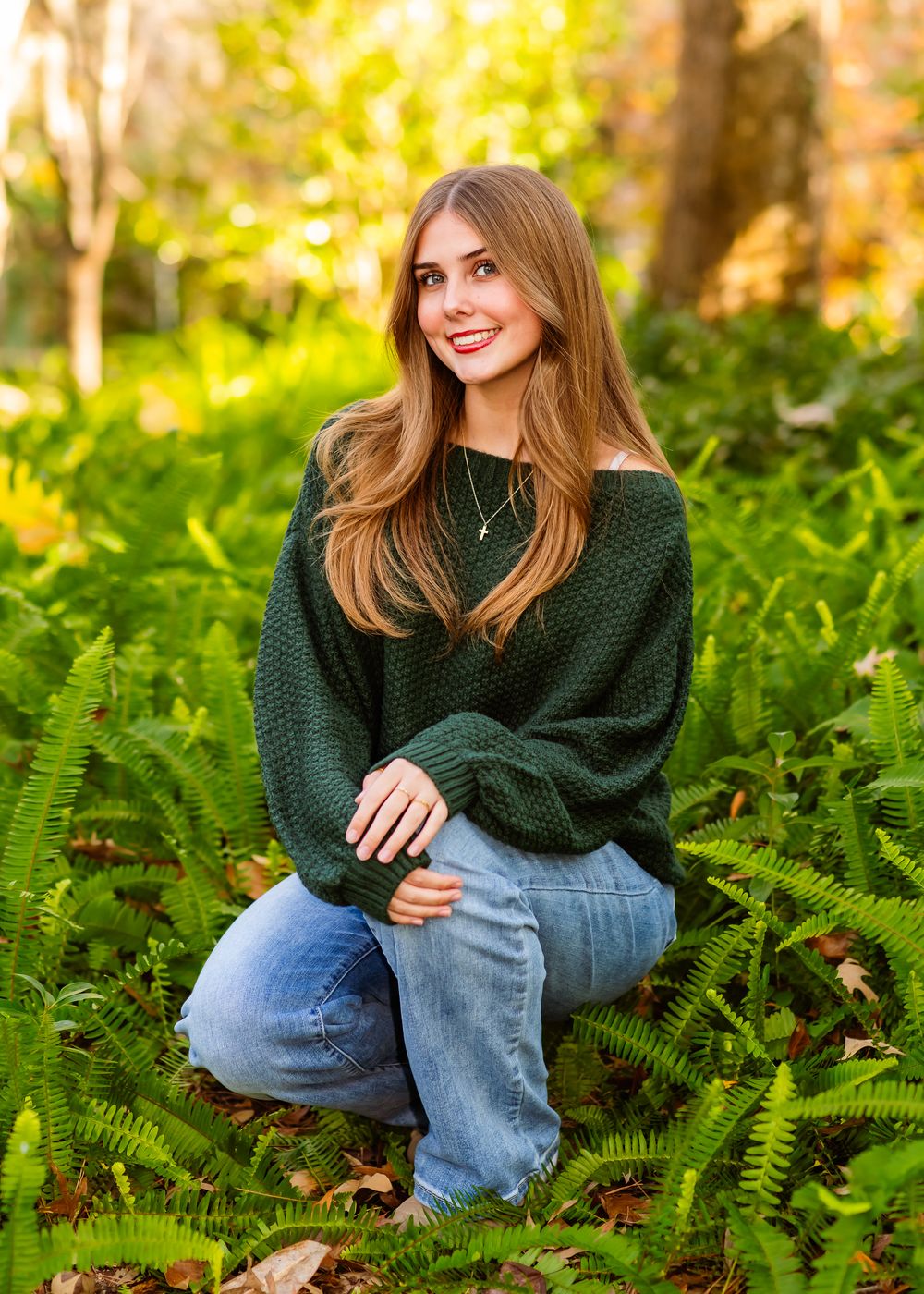 A person in a green sweater and jeans sits among lush ferns in a sunlit forest setting.
