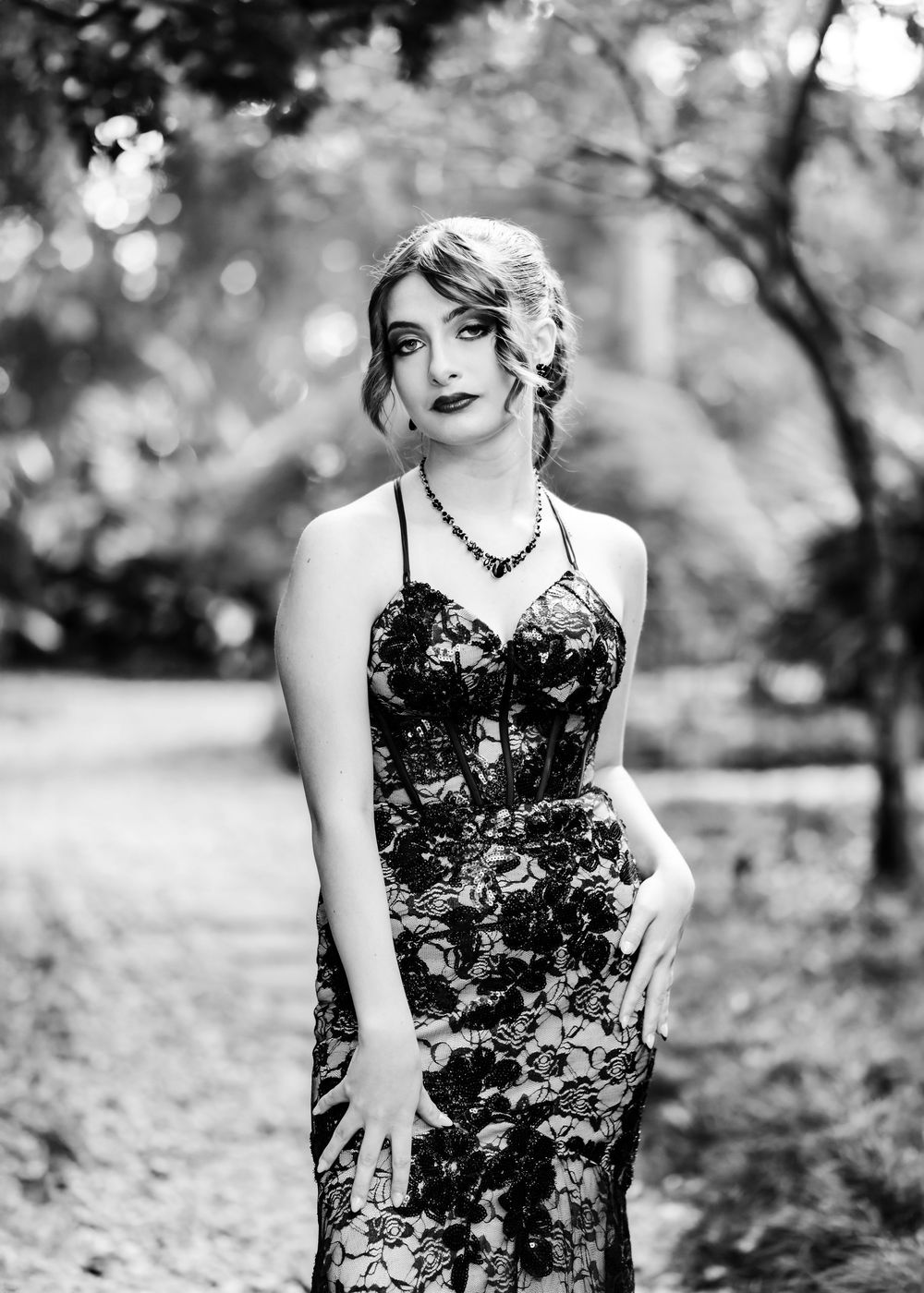Black and white portrait of a woman in a floral lace gown and necklace posing outdoors among trees.