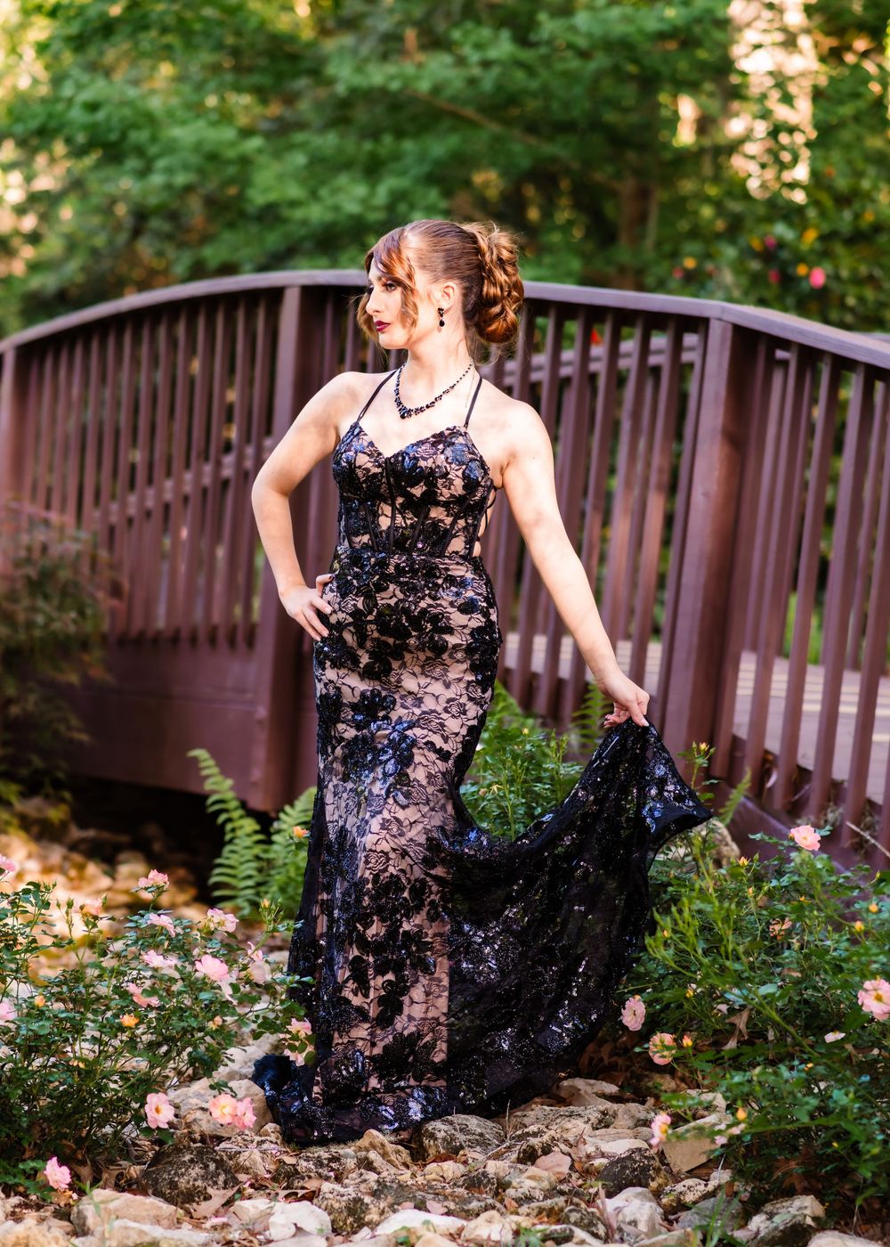 Woman in black lace gown poses gracefully on wooden bridge surrounded by lush greenery and pink roses.
