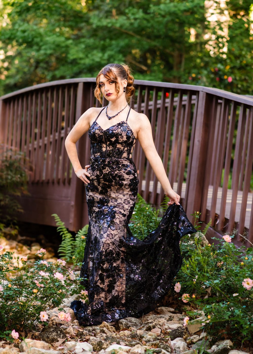 Young woman in black lace floral gown poses gracefully on a wooden bridge surrounded by lush greenery and roses.