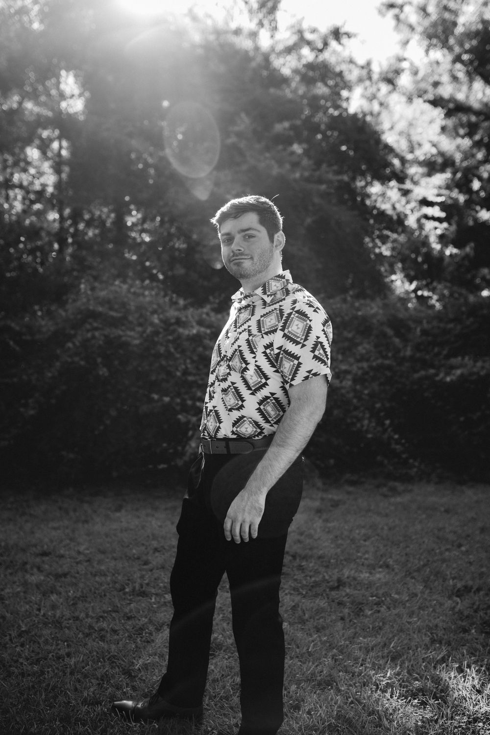 A black and white outdoor portrait showing someone wearing a patterned short-sleeve shirt and dark pants in sunlit scenery.