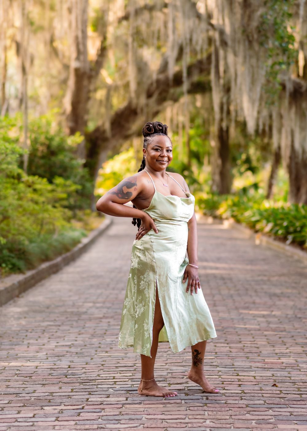 Woman in mint green dress poses on brick path surrounded by moss-draped trees in a lush garden setting.