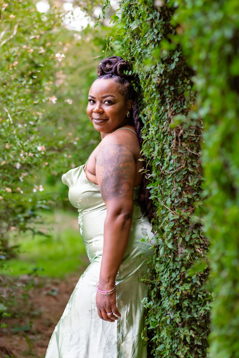 A smiling woman in a sage green gown leans against an ivy-covered pillar in a lush garden setting.