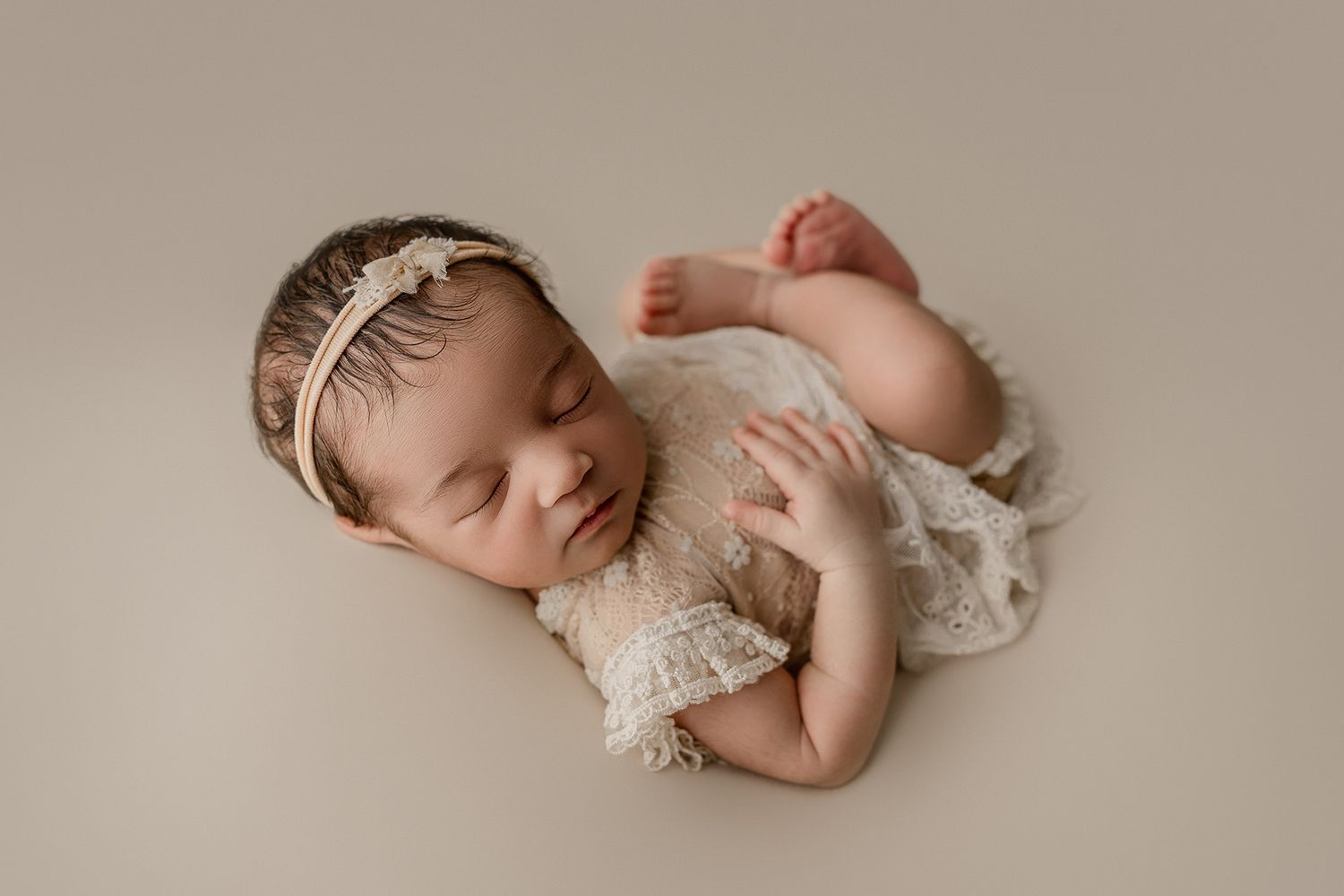 Peaceful newborn baby girl sleeping in cream lace romper and floral headband on neutral beige backdrop.