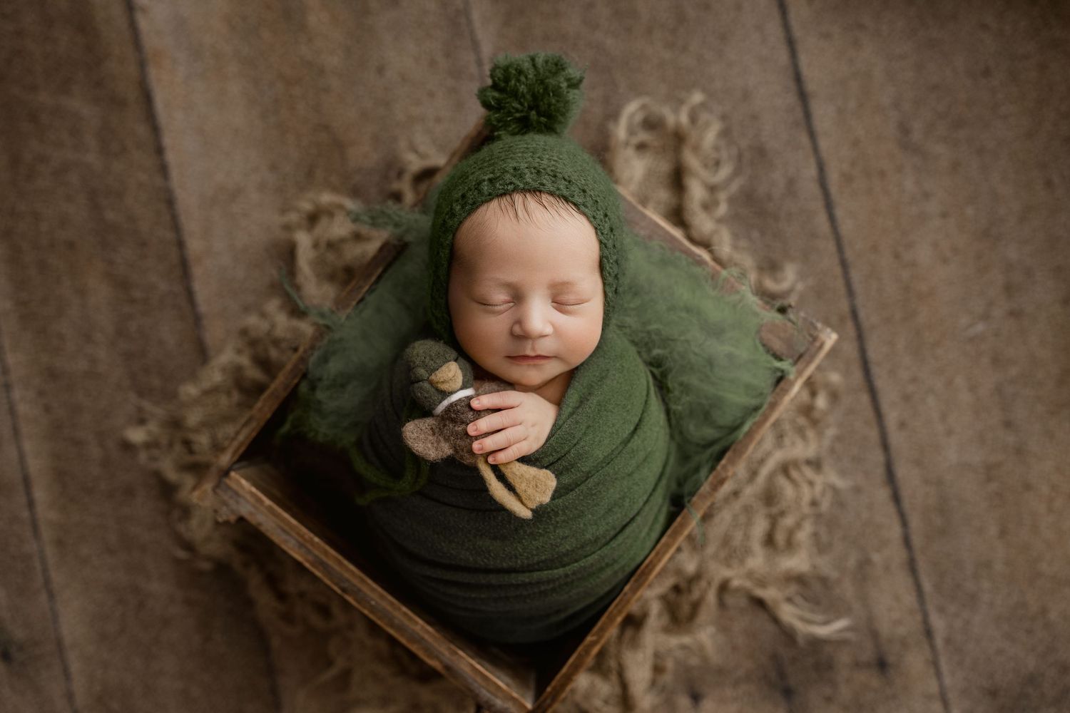 Newborn baby wrapped in green knit swaddle wearing a pom-pom bonnet, sleeping in a rustic wooden crate.