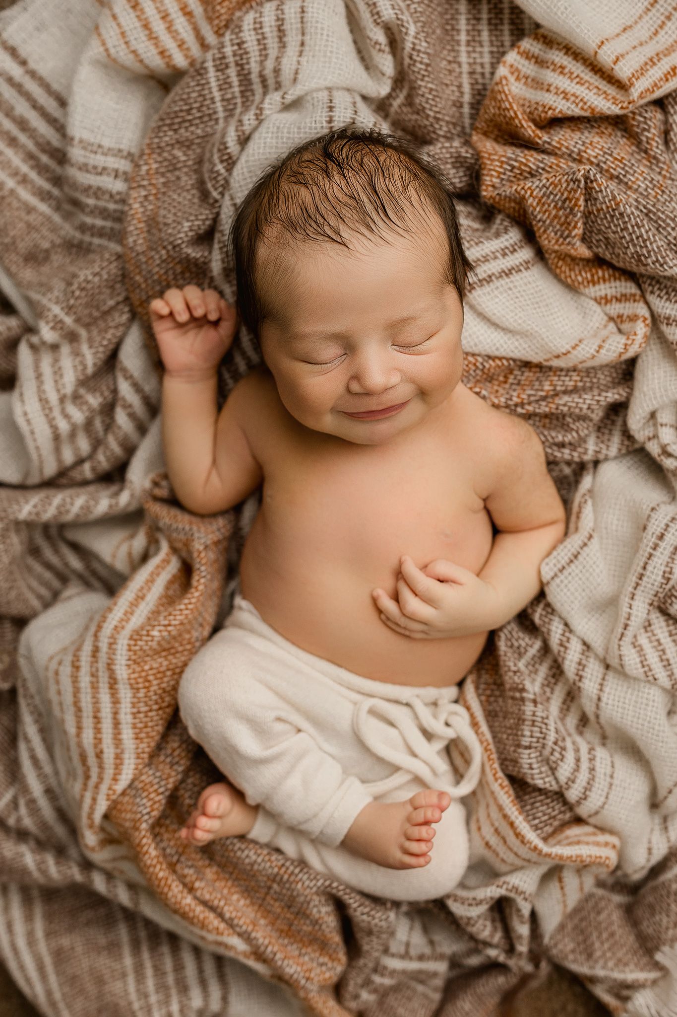 Smiling newborn baby sleeping peacefully on brown striped blankets wearing white pants.