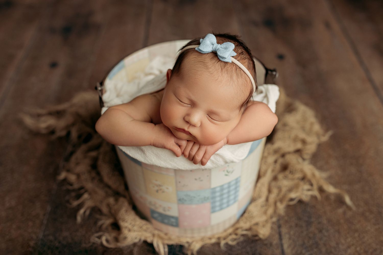 Sleeping newborn baby girl in a decorative bucket with a blue bow headband on a rustic wood background.