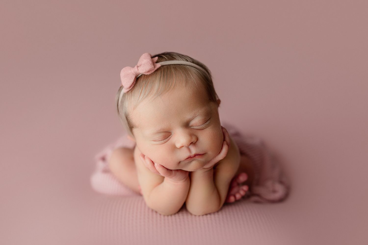 Sleeping newborn baby girl in pink outfit and bow headband posed with hands under chin on pink backdrop.