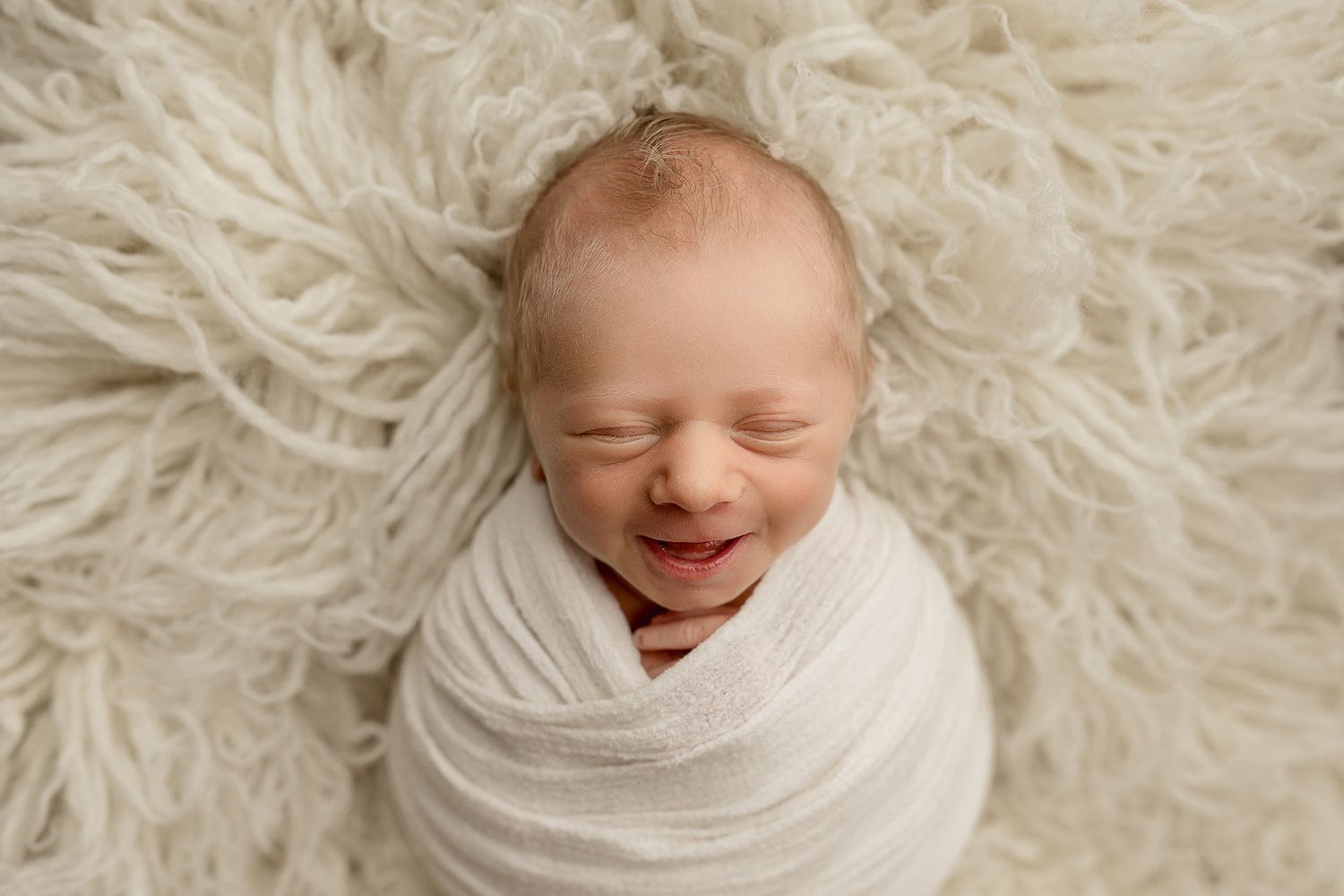 Smiling newborn baby swaddled in white wrap, peacefully sleeping on a fluffy cream fur blanket.