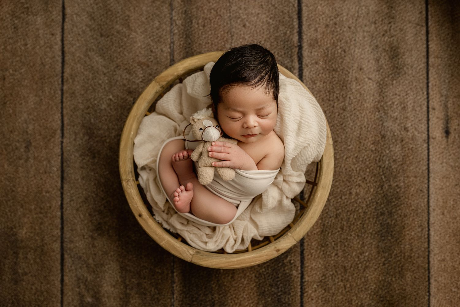 Sleeping newborn baby in wooden bowl with cream wrap holding tiny stuffed animal toy, peaceful nursery portrait.