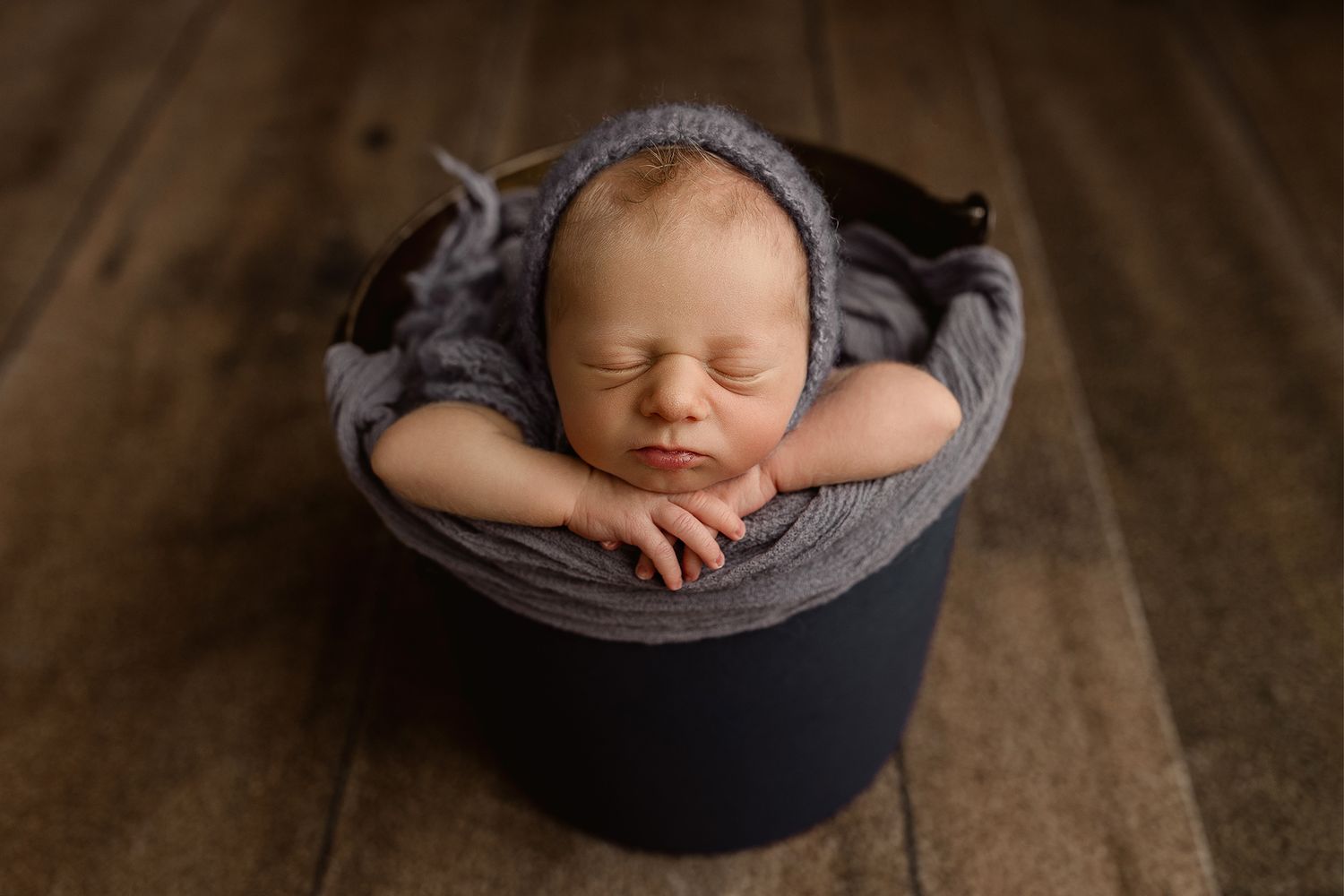 Sleeping newborn baby in gray knit bonnet posed in dark bucket wrapped in soft gray fabric on wood floor.
