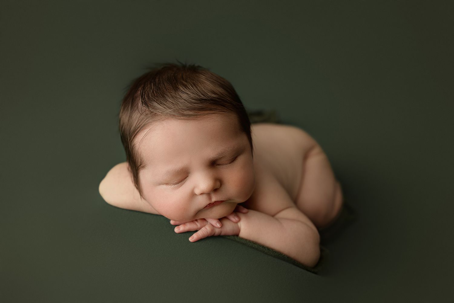 Peaceful newborn baby sleeping on green backdrop with hands tucked under chin in classic froggy pose.