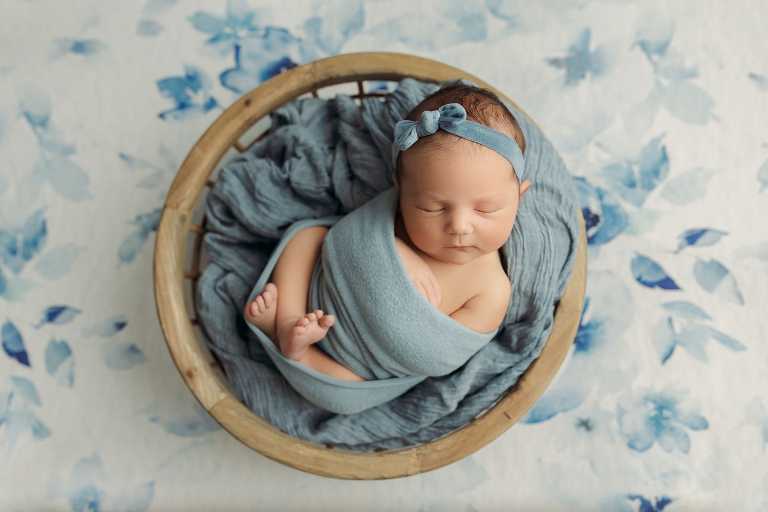 Newborn baby wrapped in blue fabric sleeping peacefully in a wooden bowl on a floral blue background.
