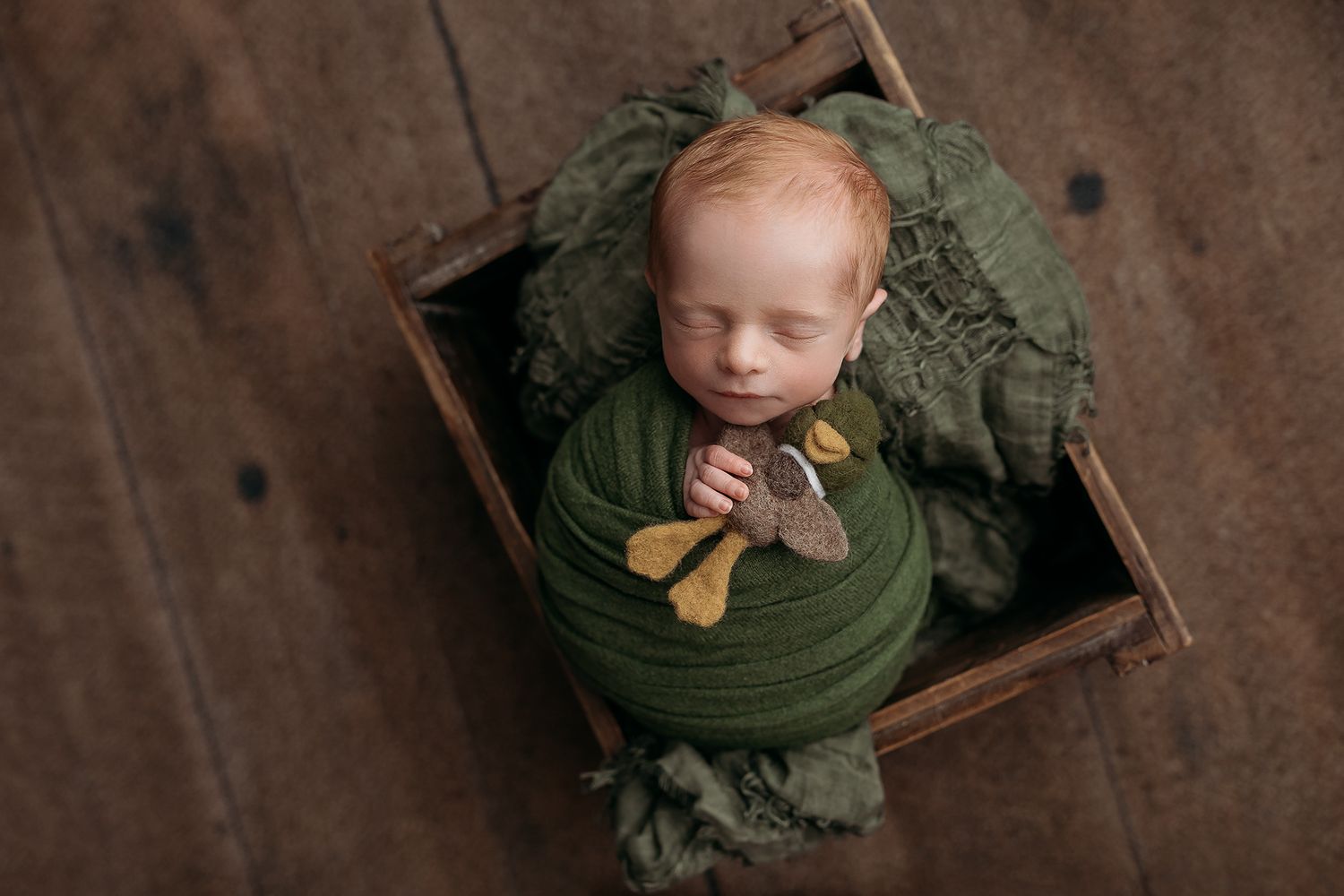 Newborn baby wrapped in green swaddle sleeping in rustic wooden crate with tiny bow tie prop.