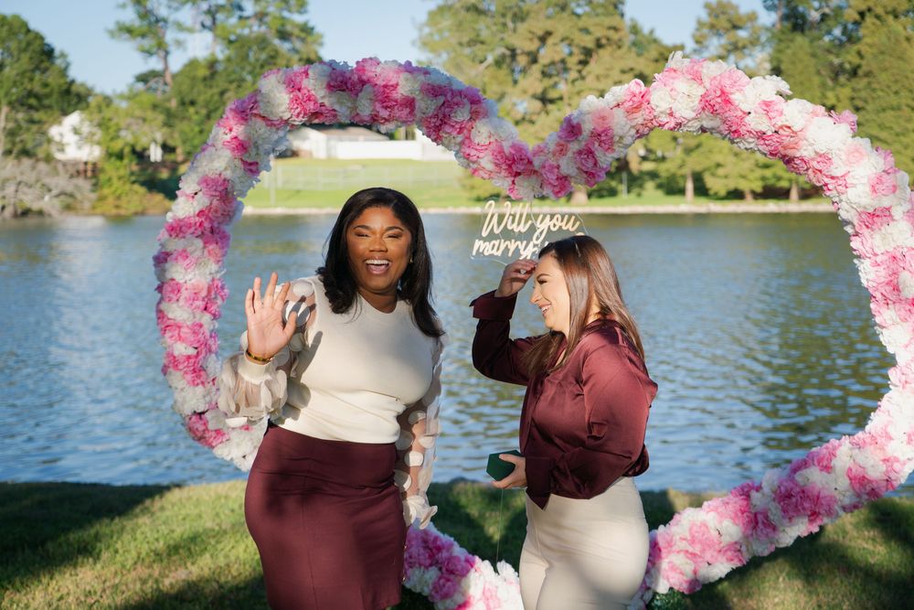 Two people pose together by a lake with pink floral decorations and trees in the background.
