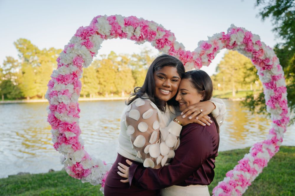 Two people embrace while standing in front of a pink floral heart arch by a scenic lake during golden hour.