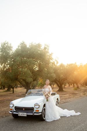 Classic white Mercedes convertible parked in an olive grove at sunset with flowing wedding dress draped over side.