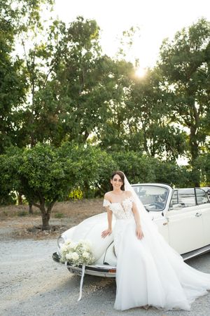 A classic white convertible car parked in an orchard during golden hour with flowing wedding dress details.