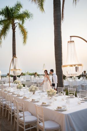 Elegant outdoor wedding reception setup with white tables, crystal chandeliers and palm trees at sunset.