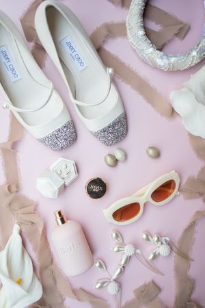 Collection of elegant white bridal shoes and accessories arranged on pink background.