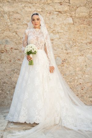 Elegant long-sleeved lace wedding gown with cathedral train against stone wall.
