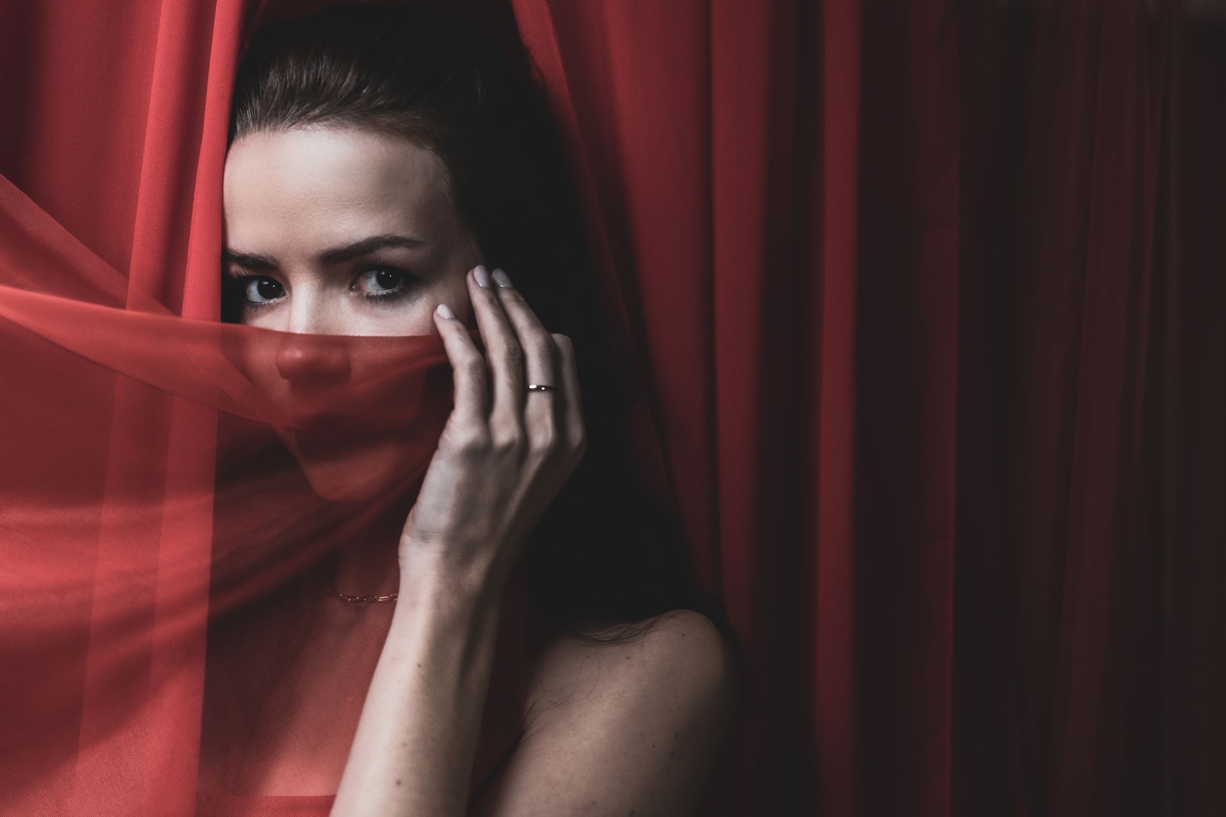 Figure in dramatic red lighting poses mysteriously behind sheer fabric creating an ethereal portrait series.