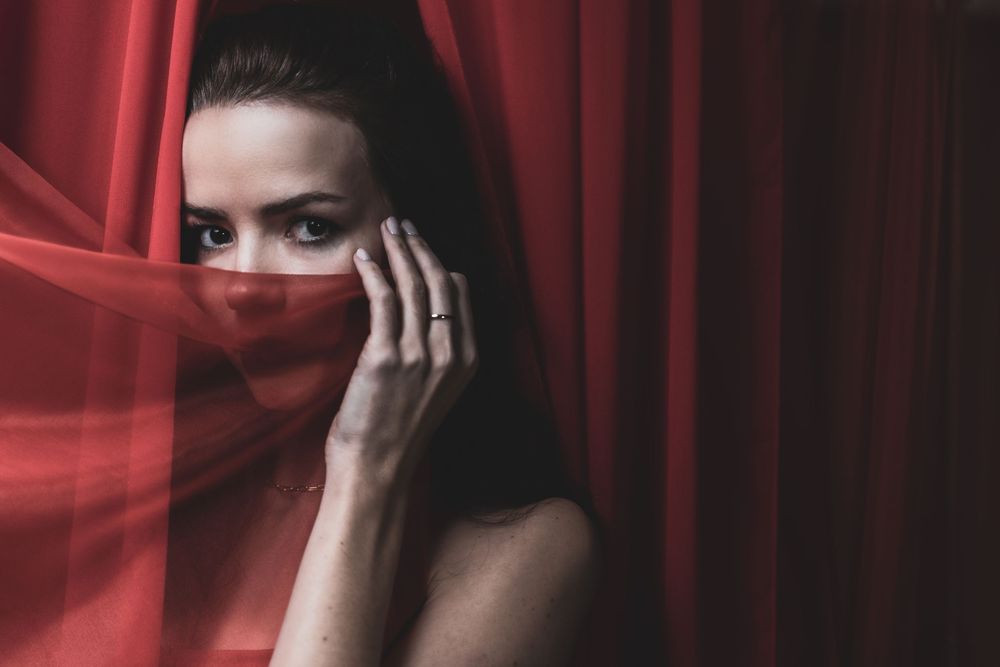 Figure in dramatic red lighting poses mysteriously behind sheer fabric creating an ethereal portrait series.