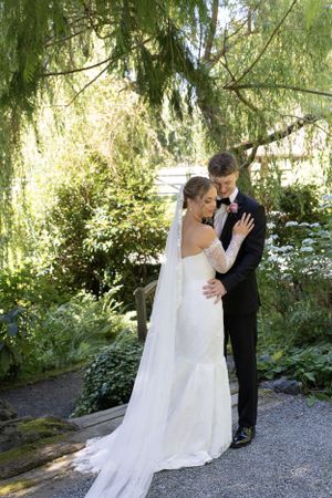 Cora Emilie Media A newlywed couple embraces on a garden path under weeping willow trees.
