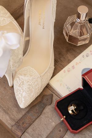 Cora Emilie Media Elegant white lace bridal shoes with perfume bottles on wooden surface.