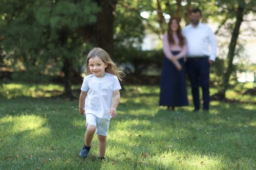 Candid lifestyle photo of a young boy running through the grass with her parents in the blurred background during an outdoor family session