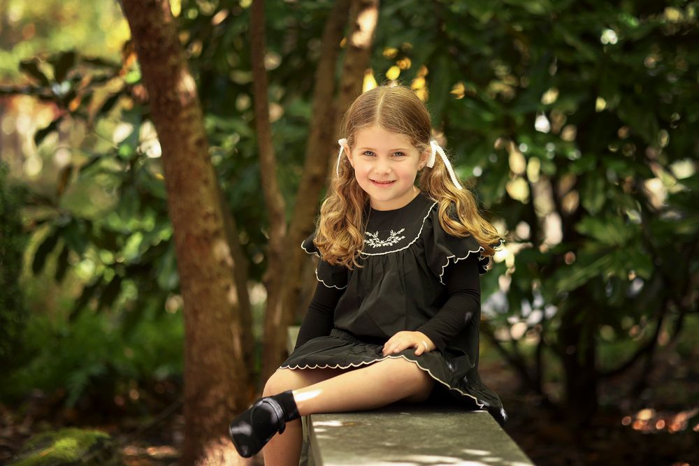 A professional outdoor portrait of a young girl with pigtails and white bows sitting on a stone bench in a lush garden in Jackson, NJ