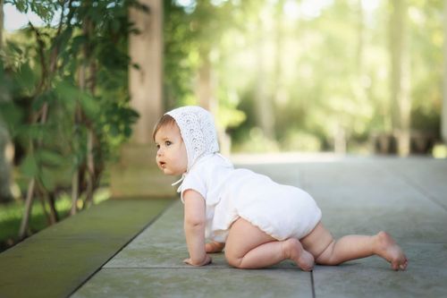 outdoor fine art candid shot of crawling infant wearing a white knot bonnet with natural greenery in the background