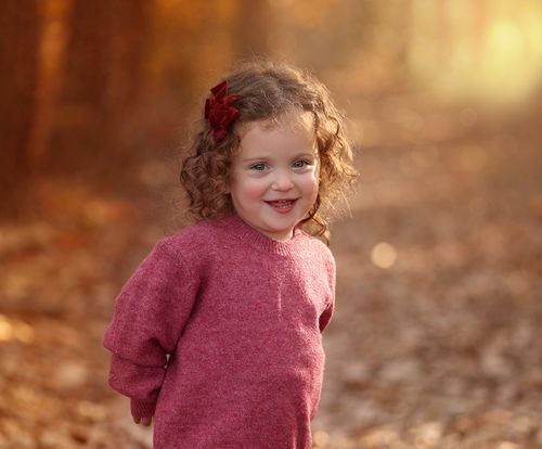 A lifestyle outdoor portrait of a young girl with curly hair and a red bow smiling on a wooded forest path during an autumn photo shoot