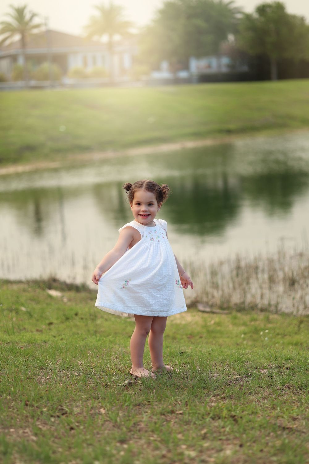 toddler girl showing off her pretty dress while standing in front of a sun drenched lake