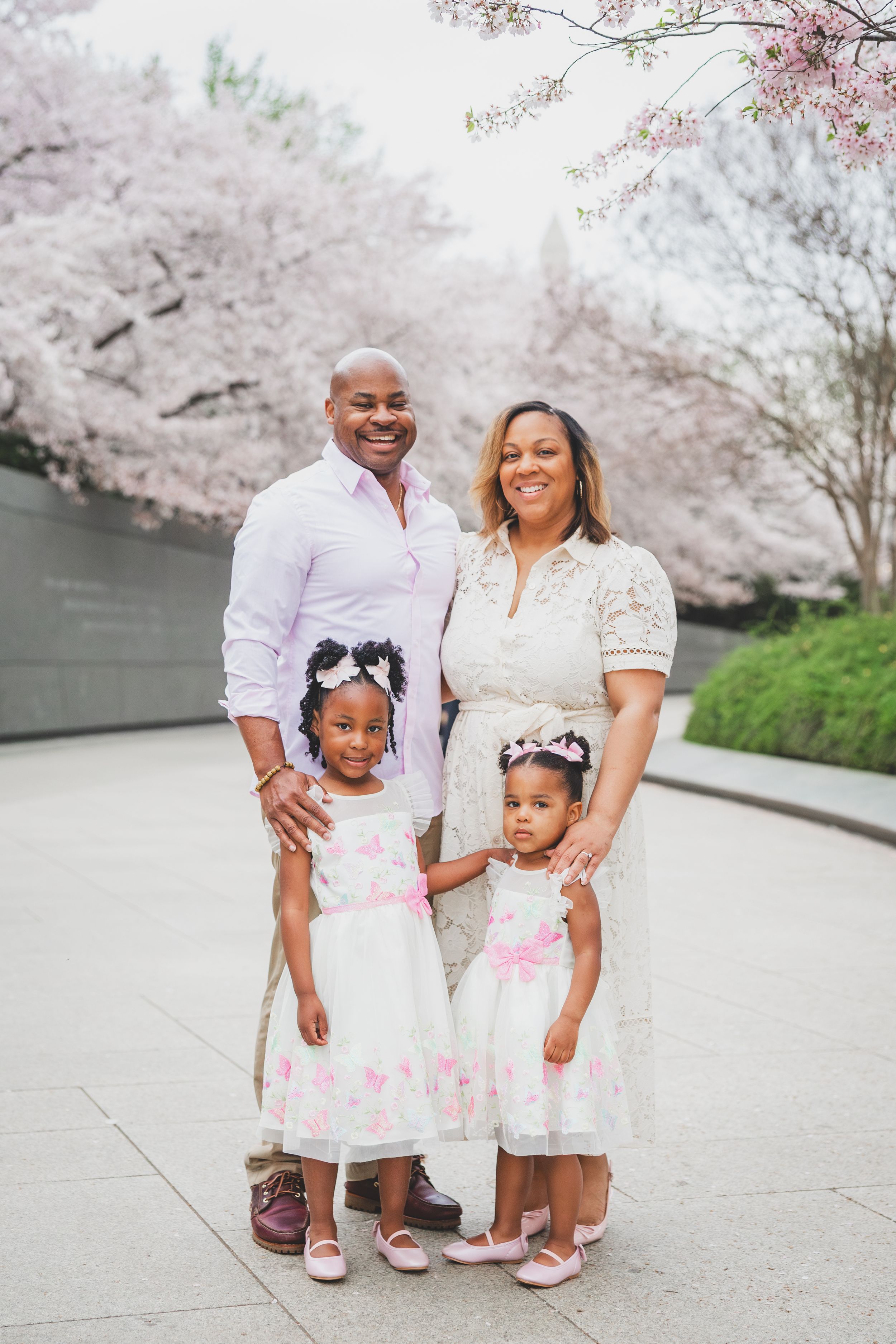 Family dressed in coordinated white outfits poses together under blooming cherry blossom trees during springtime.