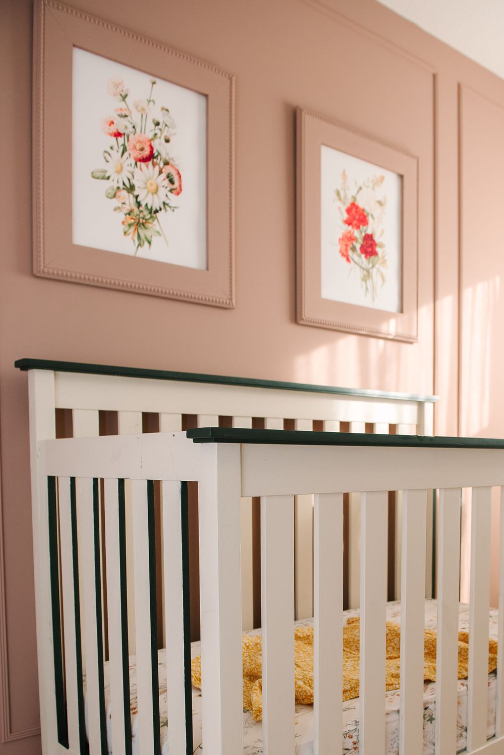 Nursery wall with pink paint and white framed floral artwork above a white crib railing.